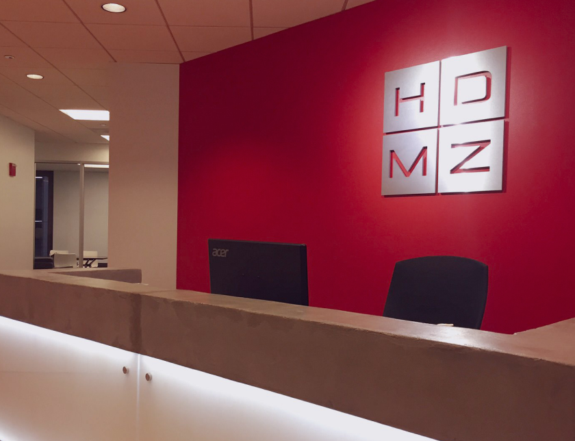 A photo of the entryway of HDMZ's home office. The HDMZ logo is featured in silver in front of a red wall.