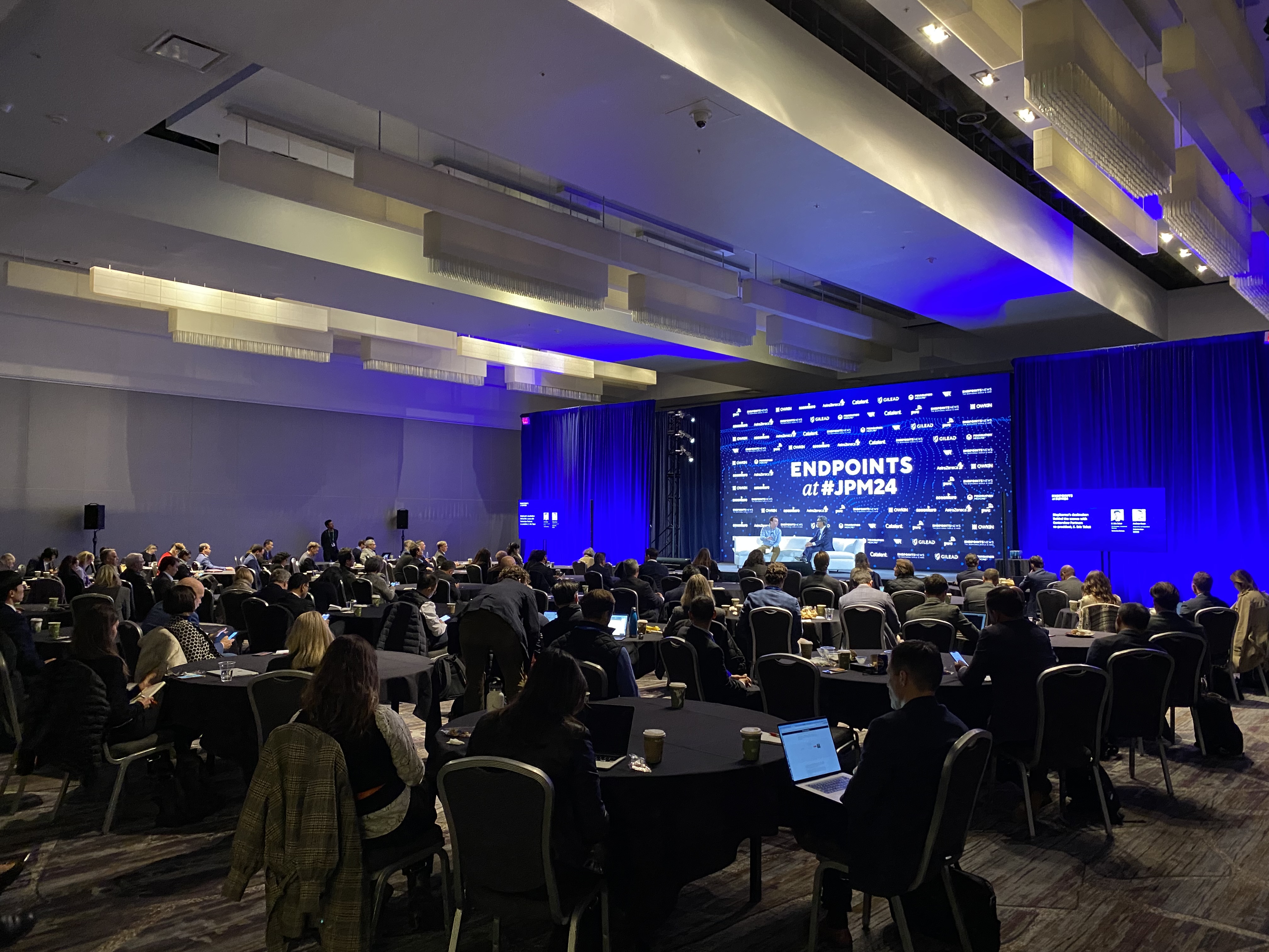 Two male speakers on stage at JPM 2024 Endpoints with audience sitting at tables